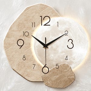 Puede incluir: Un reloj de pared con una forma de media luna de color beige con textura de piedra que se superpone a una esfera circular iluminada similar a la luna. El reloj tiene manecillas y números negros del 1 al 12. Una luz cálida brilla detrás.