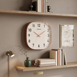 May include: A square wall clock with rounded corners, featuring a white face, black numerals, and rose gold hands. The brand name Mobpedo is printed in black on the clock face. The clock has a metallic rose gold frame and is mounted on a wall.