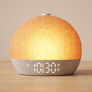 May include: A dome-shaped digital alarm clock with a textured fabric finish. The upper half glows with a warm orange light, while the lower grey base displays the time 10:30 in white digital numerals, along with AM, snooze, music, and alarm icons.