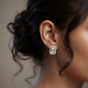 May include: A rectangular emerald-cut center stone earring set in a gold metal frame, surrounded by a halo of small round clear stones. The earring is secured to the earlobe with a post backing, featuring a classic design with metallic gold tones.