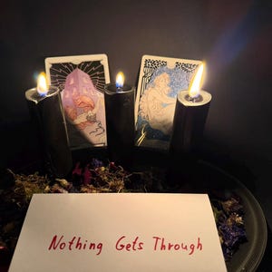 May include: Three black pillar candles with lit flames are arranged behind a white rectangular card featuring the red handwritten text Nothing Gets Through. Two illustrated tarot cards are positioned behind the candles, surrounded by dried flowers.