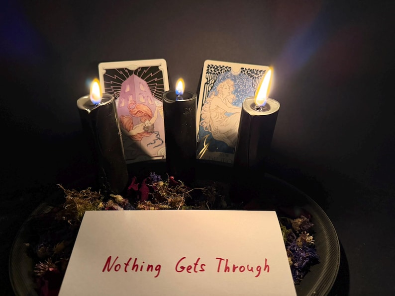 May include: Three black pillar candles with lit flames are arranged behind a white rectangular card featuring the red handwritten text Nothing Gets Through. Two illustrated tarot cards are positioned behind the candles, surrounded by dried flowers.