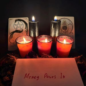 May include: Three red votive candles in glass holders and two tall black pillar candles are lit. Behind them are two tarot cards. In the foreground, a white card features the handwritten red text "Money Pours In" with dried botanical elements.