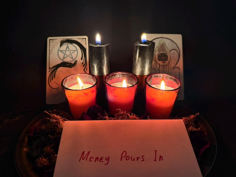 May include: Three red votive candles in glass holders and two tall black pillar candles are lit. Behind them are two tarot cards. In the foreground, a white card features the handwritten red text "Money Pours In" with dried botanical elements.