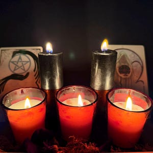 May include: Two tall black pillar candles and three red votive candles in glass holders, all with lit flames. Behind the candles are two rectangular cards, one featuring a pentacle symbol and the other an illustration of a hooded figure.