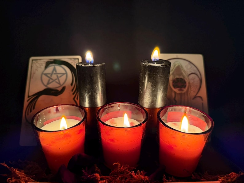 May include: Two tall black pillar candles and three red votive candles in glass holders, all with lit flames. Behind the candles are two rectangular cards, one featuring a pentacle symbol and the other an illustration of a hooded figure.