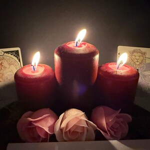 May include: Three deep red pillar candles with lit flames arranged in a row, flanked by two illustrated tarot cards featuring a couple and a lion. Three soft pink rose blossoms are positioned in front of the candles against a dark background.