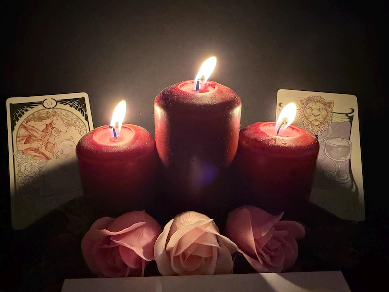 May include: Three deep red pillar candles with lit flames arranged in a row, flanked by two illustrated tarot cards featuring a couple and a lion. Three soft pink rose blossoms are positioned in front of the candles against a dark background.