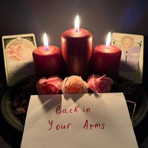 May include: Three dark red pillar candles with lit flames arranged on a black plate with three light pink artificial roses and two illustrated tarot cards. A white rectangular card in the foreground features the handwritten text Back in Your Arms.