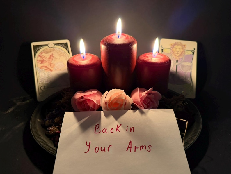 May include: Three dark red pillar candles with lit flames arranged on a black plate with three light pink artificial roses and two illustrated tarot cards. A white rectangular card in the foreground features the handwritten text Back in Your Arms.