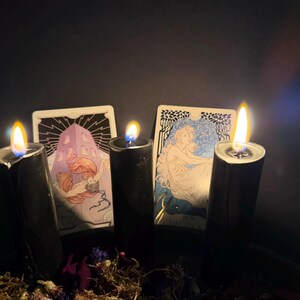May include: Three black pillar candles with lit flames are positioned in front of two tarot cards. The left card depicts a figure with long hair near a structure, while the right card shows a seated figure. Dried flowers are at the base.