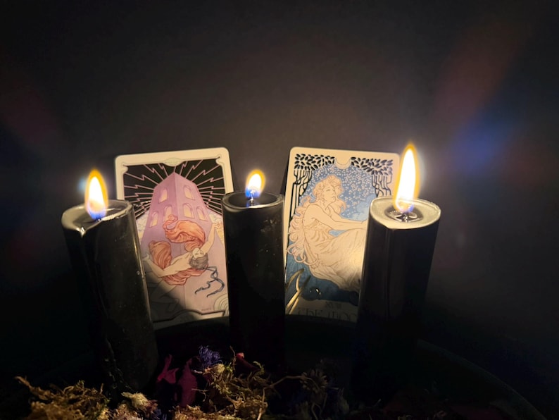 May include: Three black pillar candles with lit flames are positioned in front of two tarot cards. The left card depicts a figure with long hair near a structure, while the right card shows a seated figure. Dried flowers are at the base.