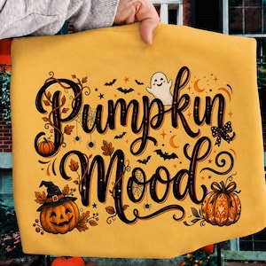 以下が含まれることがあります： マスタードイエローのトレーナーで、黒い筆記体で「Pumpkin Mood」という文字がプリントされています。デザインには、笑顔のゴースト、魔女の帽子をかぶったジャック・オー・ランタン、模様入りのカボチャ、クモ、クモの巣、コウモリ、小さな星が描かれています。