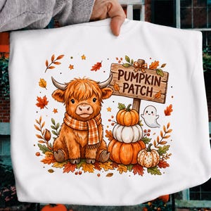 May include: A white t-shirt featuring a graphic of a fluffy brown highland cow wearing a plaid scarf, sitting next to a stack of three pumpkins and a small ghost. A wooden sign above reads PUMPKIN PATCH, surrounded by colorful autumn leaves.