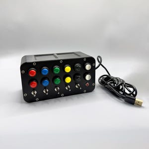 May include: A black rectangular control box featuring two rows of colourful push buttons in red, blue, green, yellow, black, and white. The device includes five metal toggle switches along the bottom edge and a black USB cable with a connector.