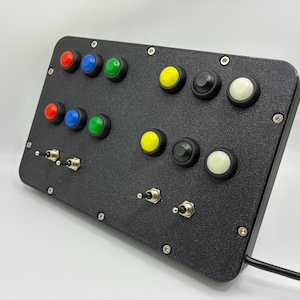 May include: A black rectangular control box featuring a textured surface with six colourful push buttons in red, blue, and green, plus six additional buttons in yellow, black, and white. Four metal toggle switches are positioned on the front panel.