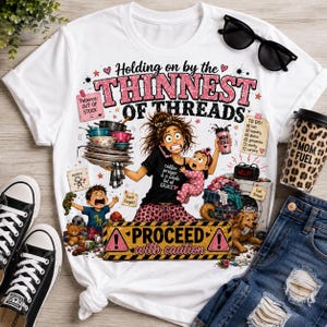 May include: A white t-shirt featuring a colorful graphic illustration of a stressed parent holding a baby, surrounded by household chaos, a stack of dishes, a laundry basket, a dog, and text that reads Holding on by the THINNEST OF THREADS.