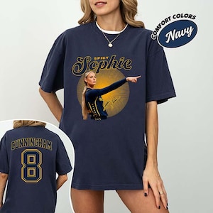 May include: A navy blue short-sleeve t-shirt featuring a graphic of a basketball player with the text Spicy Sophie above a yellow circular pattern. The back of the shirt displays the name CUNNINGHAM and the number 8 in yellow and navy blue.