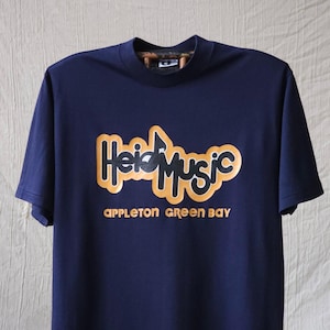 以下が含まれることがあります： 黒い文字にゴールドの縁取りで「Heid Music」、その下にゴールドの文字で「APPLETON GREEN BAY」とプリントされたネイビーの半袖Tシャツ。ハイネック仕様で、襟の内側には迷彩柄があしらわれています。