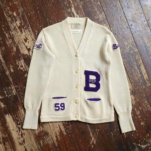 May include: A cream-colored V-neck cardigan sweater featuring purple felt patches. The front displays a large letter B with the text PEP, the number 59, and smaller patches on the shoulders. It has five light-colored buttons and ribbed cuffs.