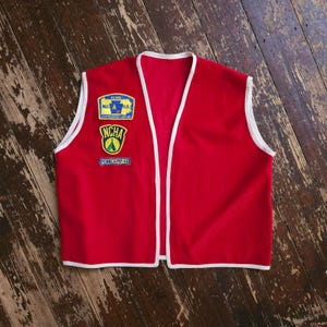 May include: A bright red sleeveless vest featuring white trim along the edges. The front right side displays three embroidered patches: a blue and yellow N.C.H.A. State Association patch, a yellow and green NCHA tent patch, and a blue Penncamp-68 patch.