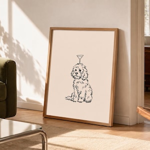 May include: A framed wall art print featuring a black line drawing of a fluffy dog sitting with a martini glass balanced on its head. The artwork is set against a solid light beige background and enclosed in a simple light wood frame.