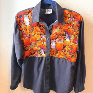 May include: A long-sleeved button-down shirt with a solid dark gray base and a contrasting panel across the chest and shoulders. The panel features a vibrant Halloween-themed print of orange pumpkins, autumn leaves, and small teddy bears in costumes.