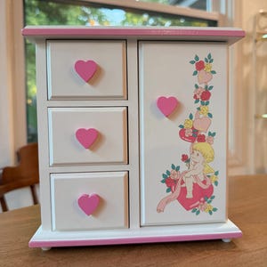 May include: A white wooden jewelry armoire featuring three drawers on the left and a single door on the right. Each drawer and the door have a pink heart-shaped knob. The door displays a graphic of a cherub sitting on a heart with roses.