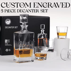May include: A 5-piece decanter set featuring a large square glass decanter with a stopper and four matching square glasses. The set is displayed in front of a black box labeled CUSTOM ENGRAVED 5 PIECE DECANTER SET. Some glasses contain amber liquid.