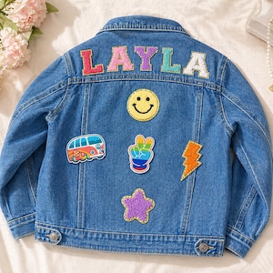 May include: A blue denim jacket featuring the name LAYLA in colourful glittery letters across the back. The jacket is decorated with several patches including a yellow smiley face, a colourful camper van, a peace sign hand, a lightning bolt, and a star.