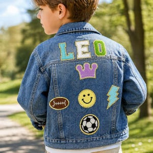 Peut inclure: Une veste en jean bleu avec le nom LEO en lettres majuscules colorées sur le haut. Sous le nom se trouvent des écussons brodés incluant une couronne violette, un ballon de rugby, un visage souriant jaune, un éclair bleu et un ballon de football.