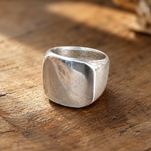May include: A solid silver signet ring featuring a smooth, polished, and slightly rounded square face. The metal surface displays a reflective, metallic sheen with subtle variations in texture, showcasing a thick, substantial band design.