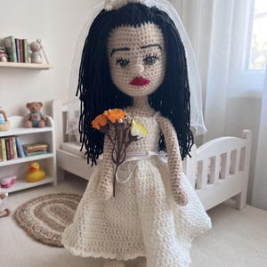 May include: A handmade crochet doll with long black yarn hair, wearing a white dress and a sheer white veil. The doll has embroidered facial features, including red lips, and holds a small bouquet of orange and yellow fabric flowers in its hands.