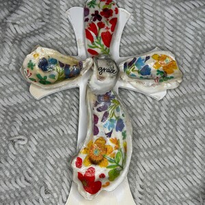 May include: A white wooden cross adorned with five oyster shells featuring colorful floral patterns. The central shell is smaller and displays the word grace in black script. A gold string is attached to the top for hanging purposes.