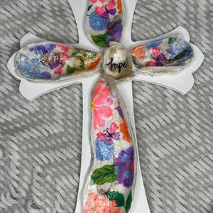 May include: A white wooden cross decorated with five oyster shells. The shells feature colorful floral and butterfly patterns, with the center shell displaying the word hope in black script. A brown twine loop is attached to the top for hanging.