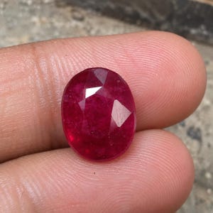 May include: A deep red, oval-shaped gemstone with a faceted surface. The stone displays a rich, saturated crimson hue and reflects light through its multiple cut planes, showcasing a translucent quality and a polished, smooth finish throughout.