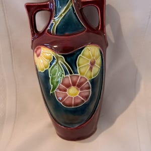 May include: A ceramic vase with a deep burgundy glaze and a dark blue central panel. The vase features two side handles and a raised relief design of three flowers in yellow, pink, and green, with green leaves and stems against the blue background.