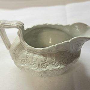 May include: A white ceramic gravy boat featuring an ornate, raised relief pattern across its exterior surface. The vessel includes a curved handle on one side and a wide, scalloped pouring spout, showcasing a detailed texture throughout the body.