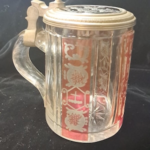 May include: A clear glass beer stein featuring vertical panels with red and white etched heraldic designs. The vessel includes a sturdy glass handle and a metallic hinged lid with a spherical finial, showcasing a detailed circular emblem on top.