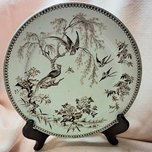 May include: A round ceramic plate featuring a dark brown transferware pattern on a light cream background. The design depicts birds, including long-tailed pheasants and swallows, perched among willow trees and blooming flowers, with a decorative border.