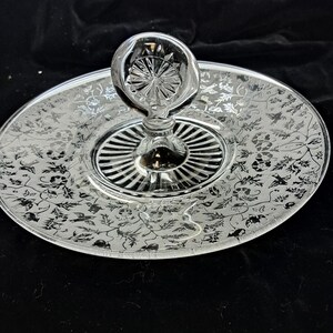 May include: A clear glass serving tray with a central handle featuring a circular, starburst-patterned top. The flat, circular plate surface is decorated with an intricate, etched silver-toned floral and bird motif across the entire rim.