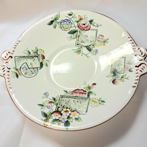 May include: A round, cream-colored ceramic serving plate with two side handles. The plate features four rectangular landscape illustrations interspersed with colorful floral clusters in shades of pink, yellow, blue, and green. The edges are trimmed in red.