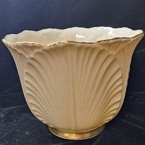 May include: A cream-colored ceramic bowl featuring a raised, vertical shell-like ribbed pattern on the exterior. The bowl has a scalloped, wavy rim and a circular base, both of which are accented with a thin, metallic gold-toned trim.