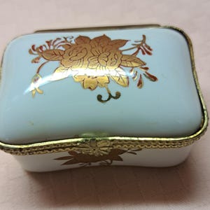 May include: A small rectangular porcelain trinket box with a white glossy finish and a gold-toned metal rim. The lid features a gold floral design, and the front side displays a gold leaf motif. The box has a hinged lid with a flower-shaped clasp.