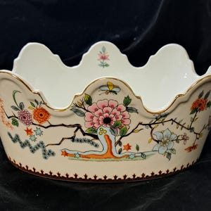May include: A cream-colored ceramic bowl with a scalloped rim and gold-tone trim. The exterior features a colorful floral pattern with pink, orange, and blue flowers, green leaves, and a small butterfly. The base has a decorative brown border.