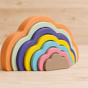 May include: A nesting toy shaped like a cloud, consisting of seven arched segments in varying sizes. The colors include orange, pale green, purple, yellow, light blue, pink, and brown, all stacked together to form a single cloud-like structure.