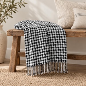 May include: A black and white houndstooth patterned throw blanket featuring a woven texture and decorative fringe along the bottom edge. The blanket is draped over a wooden bench, showcasing a classic repeating check design in high contrast colors.
