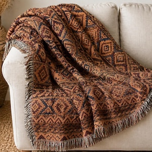 Könnte beinhalten: Eine gewebte Wohndecke mit einem sich wiederholenden geometrischen Muster in den Farben gebranntes Orange, Dunkelbraun und Anthrazit. Das Textil hat eine strukturierte Optik mit dekorativen Fransen an den Rändern und ist kunstvoll drapiert.