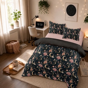 May include: A bedding set featuring a dark navy blue duvet cover and two matching pillowcases with a repeating pattern of gold crescent moons, stars, and pink floral blossoms. The set is displayed on a bed with light pink sheets and pillows.