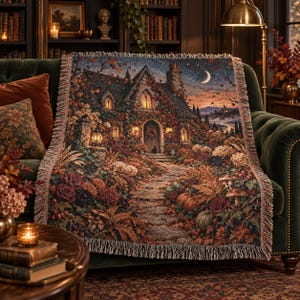 May include: A woven throw blanket featuring a detailed autumn scene of a stone cottage surrounded by fall foliage, pumpkins, and mushrooms. The design includes a stone path leading to the front door, glowing windows, and a crescent moon in the sky.